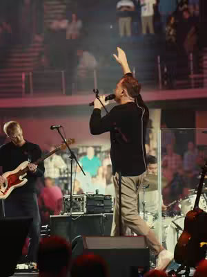 The image shows a live music performance on a stage. A central figure holds a microphone and raises their right arm, suggesting singing or worship. Two other band members include a bass guitarist on the left and a drummer in the background. The stage is well-lit, with a backdrop of blurred figures, likely an audience. The overall scene conveys a live concert environment set in a venue with a faith-based theme.