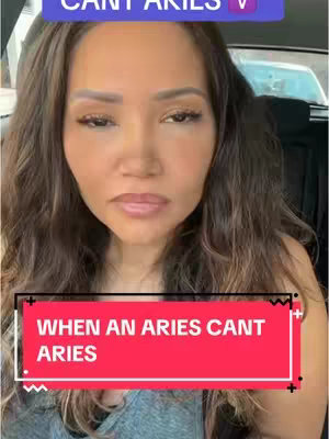 A woman is centered in a selfie frame, her brown hair flowing around her. She gazes towards the camera with a neutral expression. A rectangular text overlay at the bottom in red with white text reads: "WHEN AN ARIES CANT ARIES". The background appears to be inside a vehicle with the window visible on the right side.