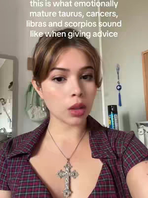 A woman with fair skin and light brown hair is the central focus. She is wearing a red and blue plaid shirt and a silver necklace with a cross pendant. The background includes a mirror and additional decor.