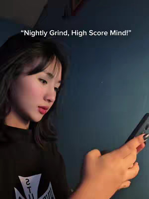 A young woman is holding a smartphone in her right hand, looking down at it with a focused expression. The background features a blue wall. Overlayed text at the top reads, Nightly Grind, High Score Mind!