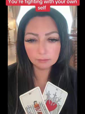A woman with long dark hair and heavy makeup is centered in frame, holding two tarot cards close to the camera. A red banner at the top of the image reads 'You're fighting with your own self'. The background is blurred, suggesting an indoor setting with curtains or drapes.