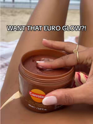 The cover image shows a close-up view of a person holding a jar that they are about to open while sitting near a beach. The person is using their finger to touch the contents of the jar, a dark brown gel. The fingers have light pink nail polish and are adorned with two gold rings. The person's legs are partially visible, and they are sitting on sandy beach. The background contains the ocean, and the sky, suggesting a sunny day. The text overlay reads "WANT THAT EURO GLOW?!"