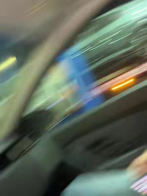 The image is heavily blurred with streaks of light and dark colors. A blurred dark shape, possibly a person, is on the left. The scene seems to be inside a vehicle, with light trails suggesting movement
