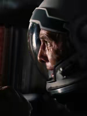 The image is a close-up shot of an astronaut's face inside a helmet. The face is weathered and appears to be in a state of contemplation or intense focus. The helmet's visor reflects the interior setting, which is dark and possibly metallic, creating a sense of enclosed space. The lighting emphasizes the astronaut's face and the details of the helmet. The overall composition is focused on the human element within the futuristic context.
