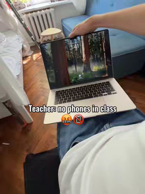 A person's lap with a MacBook Pro on it. The screen displays forest imagery. The person is wearing a white shirt and blue jeans. The room has wooden floors, a radiator and a blue sofa can be seen in the background. The text overlay reads "Teacher: no phones in class" with angry and no cell phone emojis.