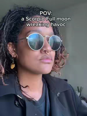 A person with curly hair and sunglasses looks to the side. Text overlay saying 'POV: a Scorpio Full moon wreaking havoc'.