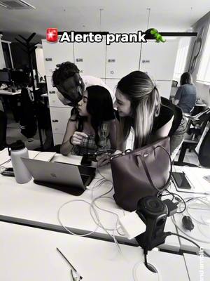 A black and white image captures three people in an office setting. A man is embracing a woman from behind, kissing her cheek, while another woman stands to the side, looking towards them. The image conveys a sense of surprise or a lighthearted moment. The background includes office furniture and equipment is present.