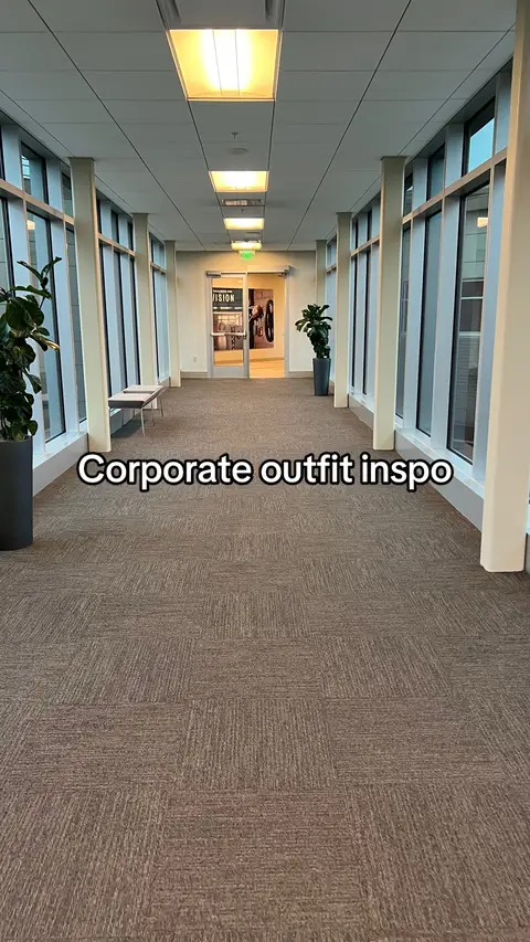 An interior shot of a long hallway. The hallway is lined with large windows on one side and a series of rectangular ceiling lights down the center. The floor is covered in a patterned brown carpet, and there are potted plants. The text overlay "Corporate outfit inspo" is visible.