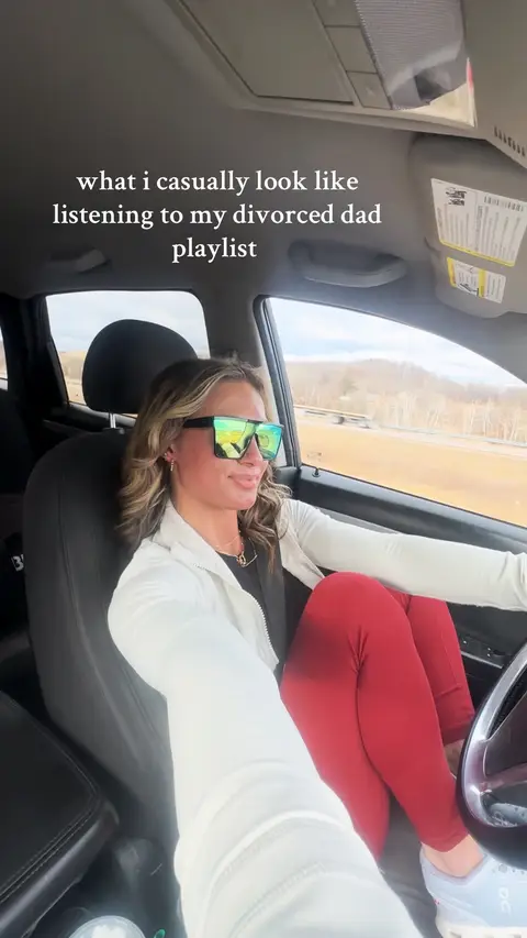 The image captures a woman in a car, viewed from a slightly low angle. She's wearing large, reflective sunglasses, a white zip-up jacket, and red leggings. The car's interior and the road outside are visible, with text overlayed on the image. The overall composition is casual and focused on the woman in a candid way.