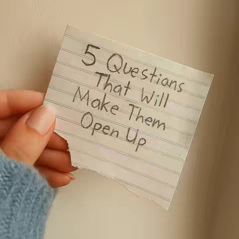 A hand holding a torn piece of lined paper with the text "5 Questions That Will Make Them Open Up" written on it in black marker. The background is blurred and appears to be a light-colored surface.