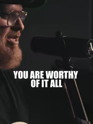 A man wearing glasses and a hat is shown from the side, singing into a microphone. Text overlayed on the image reads 'YOU ARE WORTHY OF IT ALL'. The background is dark, with soft lighting.