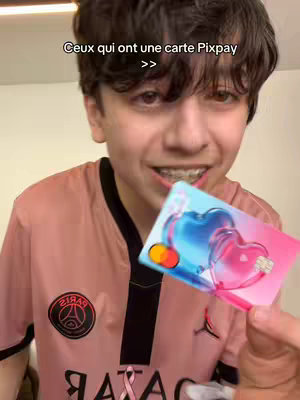 A young man with dark hair and braces holds up a credit card with a heart design, smiling at the camera.