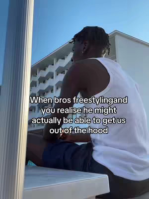 A man in a white tank top sits on a surface outdoors, looking off to the side. The background includes a building and clear blue sky. Text overlayed on the image reads, 'When bros freestyling and you realise he might actually be able to get us out of the hood'.