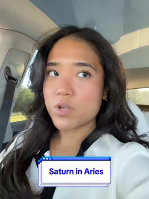 A woman is seated in a car, angled slightly towards the camera, looking off to the side with a thoughtful expression. The image is framed from a close perspective, focusing on her face, the car's interior, and a sign in the lower section.