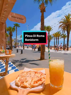 The image captures a vibrant, sunny scene at Pinsa Di Roma restaurant in Benidorm. In the foreground, a wooden table with a light orange surface holds a pizza on a wooden board and a tall glass of orange-colored drink with a straw. The background features a beach scene with people strolling along a walkway lined with palm trees under a bright blue sky. A text overlay in a color block banner reads 'Pinsa Di Roma Benidorm'. A sign for 'Pinsa Di Roma' hangs on the left.