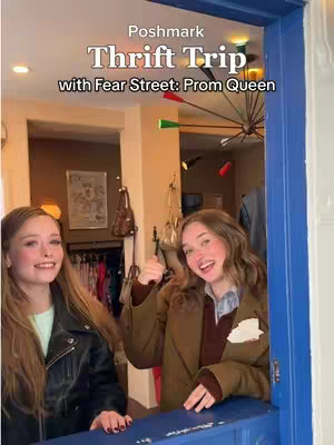 Two young women stand in a doorway of a shop, with the text overlayed. One woman on the left has long blonde hair and is wearing a black leather jacket with a white t-shirt. The other woman, on the right, has wavy brown hair and is wearing a brown coat. They are both smiling and positioned near a blue doorway with a window.