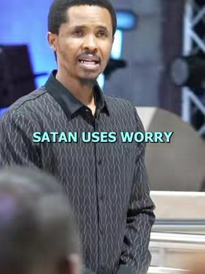 A man in a black patterned shirt stands in a bright room, speaking. Text overlays the image 'SATAN USES WORRY'. The environment hints at a stage or studio setting, possibly a place of worship or public speaking venue. The lighting is bright, and the composition focuses on the man, with a blurred background.