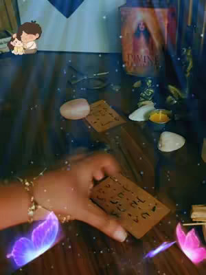 A hand wearing a gold bracelet is holding up a dark brown card with some text and hieroglyphic symbols. The hand is surrounded by colorful glitter and purple neon butterfly stickers. In the background, there is a wooden table with scattered crystals, a candle, and a small cartoon of a couple hugging.