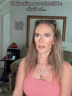 A woman with blonde hair and makeup is looking at the camera with a surprised expression. The image has text overlayed at the top: 'Zodiac Signs you should be afraid of...'. She is wearing a pink top and necklaces, and sunglasses are on top of her head. The background shows a glimpse of a room.