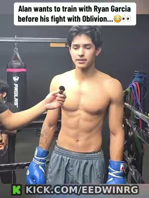 A young, shirtless man wearing boxing gloves stands in a boxing ring, with a man holding a microphone addressing him. The background has boxing equipment.