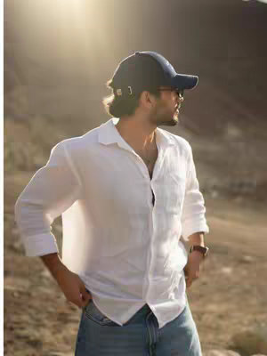 A man wearing a baseball cap, sunglasses and a white linen shirt stands outdoors in a sunlit, natural setting with mountains in the blurred background