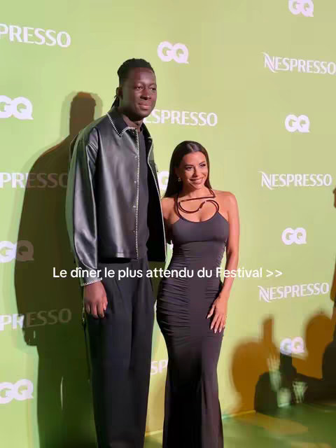 A photo taken at an event, featuring a man and a woman posing in front of a backdrop. The backdrop is green with white logos. The man is tall with dark skin, wearing a black jacket. The woman, wearing a black dress, is also smiling.