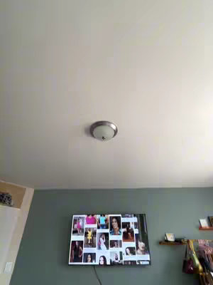 The image shows a room from a low angle, focusing on the ceiling and a portion of the wall. The ceiling, painted white, dominates the upper portion of the frame. A circular ceiling light fixture is centered, appearing to be made of metal with a glass shade. Below, a section of a gray-green wall is visible. A television screen is mounted on the wall, displaying a collage of images, likely of people. The scene gives a sense of an interior space with a focus on the ceiling.
