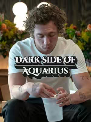 A man is sitting, with the title text overlayed. The man has curly brown hair and fair skin, and is wearing a white t-shirt. He is holding a clear plastic cup and looking down, seemingly interacting with something inside. He is sitting in front of a background of partially visible lights and plants, suggesting an interior setting.
