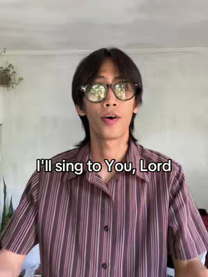 A medium shot of a young man with short black hair, wearing eyeglasses, and a vertical striped shirt looking directly at the camera with a slightly open mouth. Above 'I'll sing to you Lord'. The background is a plain wall and ceiling.