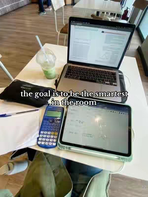 A top-down shot of a white table in a cafe. On the table, there's a laptop open with a document on the screen, next to a tablet displaying what seems like math or calculus problems. A blue calculator, a pen, and a notepad complete the study setup, and a plastic cup with a green drink and straw is there next to them. The text overlayed says "the goal is to be the smartest in the room".