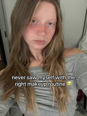 A woman with long wavy blonde hair looks directly at the camera. She is wearing a gray off-the-shoulder shirt. The background is a plain white wall with a door frame visible on the left. Text overlayed on the image reads: ''never saw myself with the right makeup routine''