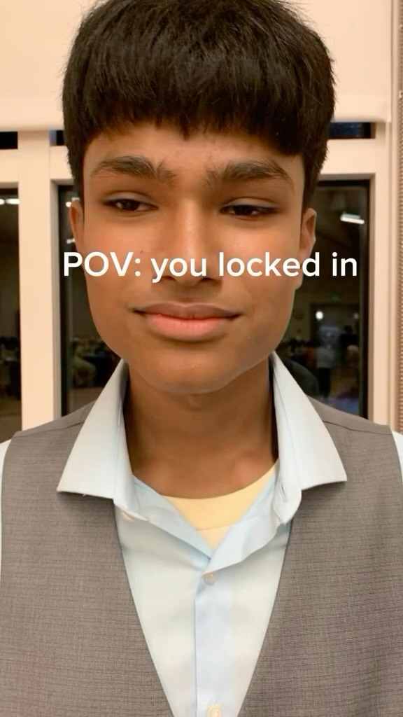 A young man with dark hair and light skin is centered in the frame. He is wearing a light blue collared shirt under a gray vest. A text overlay reads 'POV: you locked in' in white font.