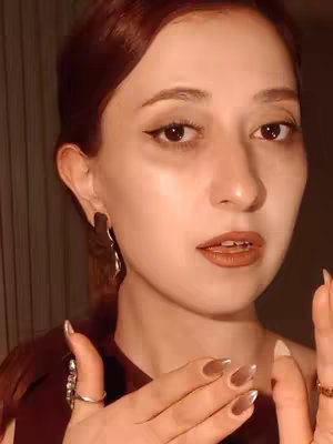 A close-up shot of a woman looking at the camera, with her hands up. She has light skin, brown hair pulled back, and is wearing a dark red top. She has makeup on, including eyeliner and lipstick. She is also wearing earrings and rings. Her nails are painted with a metallic finish.
