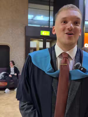 A man in a graduation gown smiling directly at the camera. He is wearing a formal suit with a white shirt, a burgundy tie, and a graduation gown. His hair is short and blond. The background is a blurred indoor setting, possibly a university or similar institution, with neutral colors and some other blurred figures.