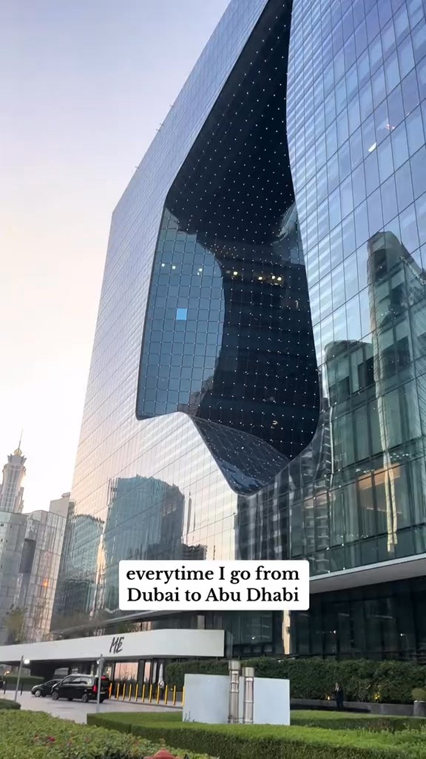 The image displays the exterior of a modern, sleek building with a unique architectural design. The building is primarily glass and reflective, with a central section carved out in an angular or wave-like shape, creating an intriguing visual element. The sky is visible above the building, suggesting a clear day. The bottom of the image includes some greenery.