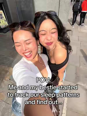 Two young women are taking a selfie outdoors, smiling and looking at the camera. The woman on the left is wearing a white top. The woman on the right is wearing a black top. Both are smiling and have sunglasses on their heads.