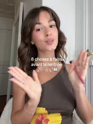 The cover image features a young woman with shoulder-length brown hair and bangs, wearing a brown top with a yellow floral design. She is positioned in the center of the frame with her hands raised, as if presenting something. Overlay text is visible above her. The background includes a white door and a glimpse of a white-painted wall.