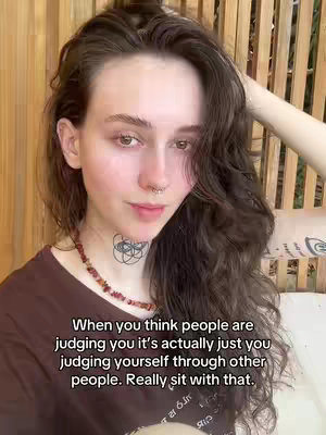 A young woman with long, wavy brown hair and a nose ring is the primary subject of the image. She's framed from the chest up, with a soft, natural lighting that highlights her features. A subtle necklace, a tattoo on her neck, and a tattoo on her arm.
