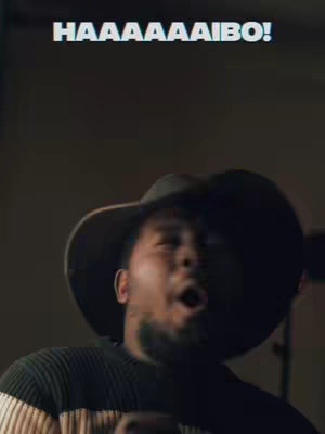 A medium shot of a Black man looking up with an open mouth as if singing. He wears a black hat that casts a shadow over his eyes, a black and white striped sweater, and has a goatee. The background is a blurred gradient, and the text 'HAAAAAAIBO!' is above his head.