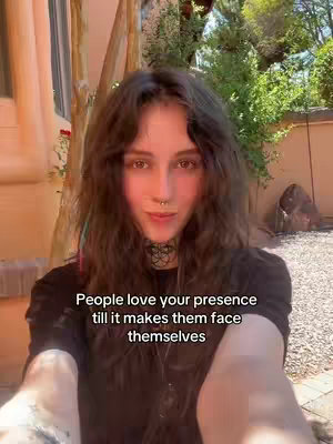 A selfie-style video captures a person with long, wavy brown hair, a nose ring, and a black choker, set against a blurred outdoor backdrop. The person looks toward the camera with a focused expression. The text overlay reads, "People love your presence till it makes them face themselves."