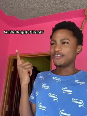 A young man in a blue Puma t-shirt with a white pattern, points with his right index finger towards the upper left of the frame. The background includes a pink wall and a door frame, and text is overlayed at the top left of the frame.
