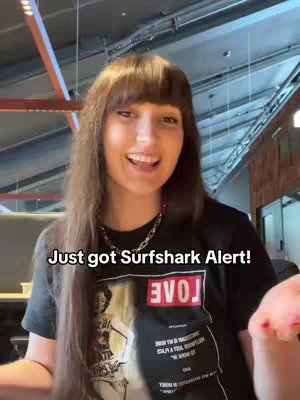 A woman with long brown hair and bangs is smiling at the camera in a brightly lit room. The text 'Just got Surfshark Alert!' is overlaid at the bottom of the frame.  She is wearing a black shirt with the word 'VOLJ' visible on the front.