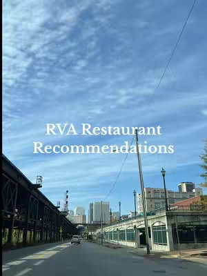 An urban street scene captured from a driving perspective. The foreground shows a road lined with a long, elevated structure on the left and a set of windows on the right. The background features city buildings and a sky with scattered clouds. The hook, overlayed in white sans-serif text, reads RVA Restaurant Recommendations.