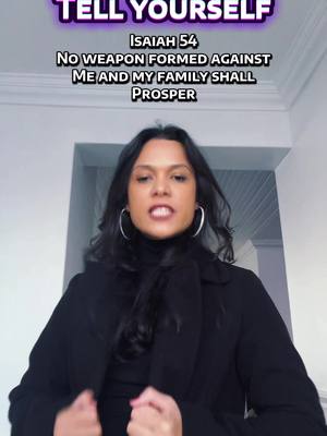 A woman with long black hair and hoop earrings is centered in the frame, wearing a black turtleneck and coat, against a light-colored wall and doorway. Text overlays appear at the top of the image.
