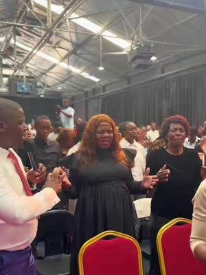 An interior church scene with an audience of people, focusing on a woman in the center. She is wearing a black dress, has her arms out, and appears to be speaking, with others around her also raising their hands. The lighting is bright, and the background includes the building's interior and additional people.