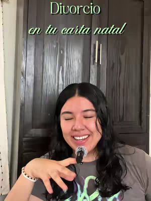 A woman with dark hair and a gray shirt is holding a microphone and smiling. Above her head, text in green letters reads "Divorcio en tu carta natal". A black wooden door serves as the background.