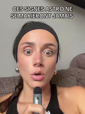A woman with large, expressive eyes and a surprised expression. She has dark hair pulled back with a black headband and is wearing hoop earrings. She holds a microphone close to her mouth, suggesting dialogue or commentary. Behind her is blurred, likely a living room setting.