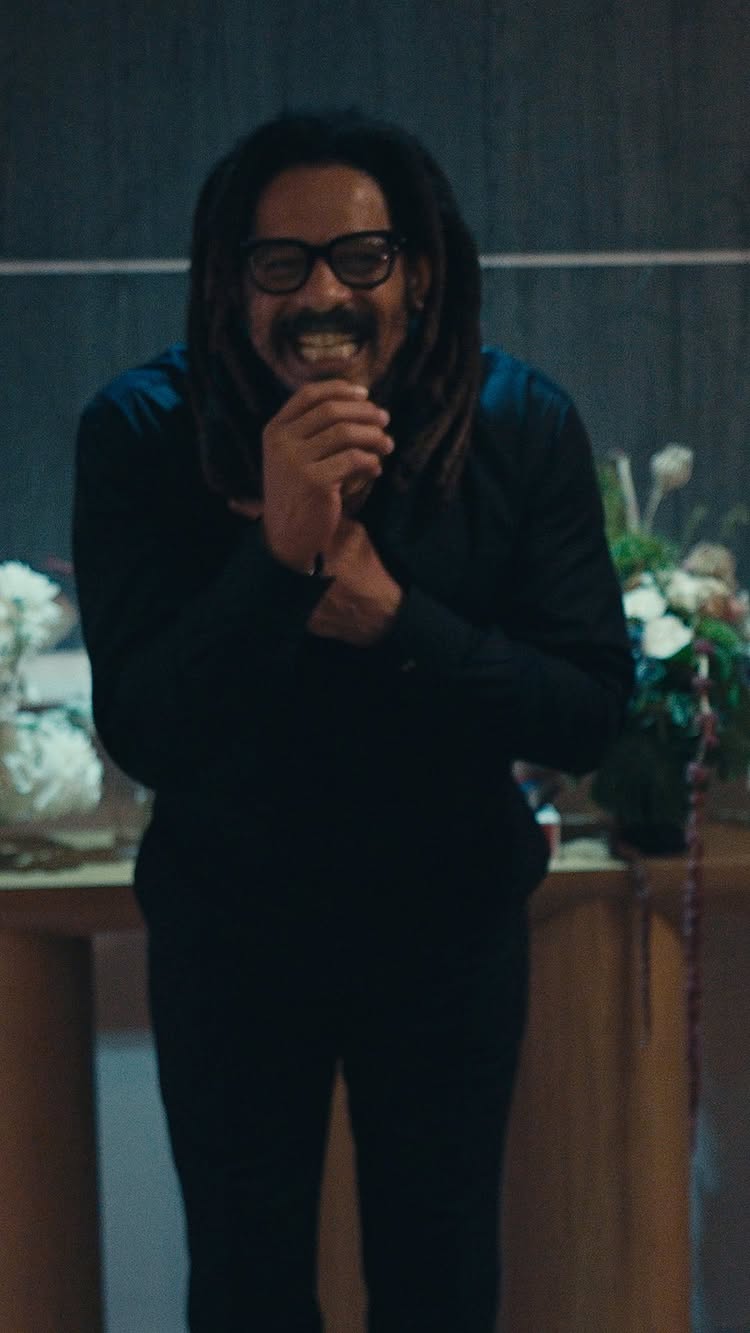 The cover image features a man wearing glasses and a mustache, smiling broadly with his hands clasped in front of his chest. He appears to be in a casual setting, with a dark background and some flowers and decorative elements visible.