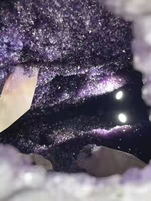 An interior shot of an amethyst geode, featuring a deep purple crystalline structure. The geode's interior is lined with tightly packed, small amethyst crystals, creating a textured surface. A dark crevice in the center may contain reflected light and the geode is framed with an irregular opening on the top.