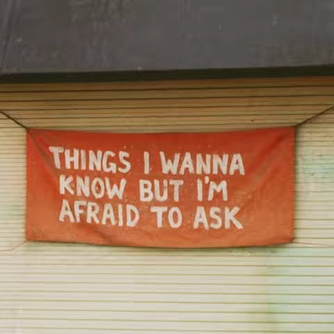 An orange banner with white text hangs against a backdrop of a light beige, horizontally-lined surface, likely the side of a building. Above the banner, there's a dark gray, possibly painted, surface that forms a partial border.